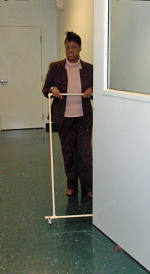 Three photos show the woman approaching the same door but holding an AMD instead of a cane.  The AMD is in the shape of a rectangle with wheels on the bottom, and she is pushing it like one would push a lawnmower.